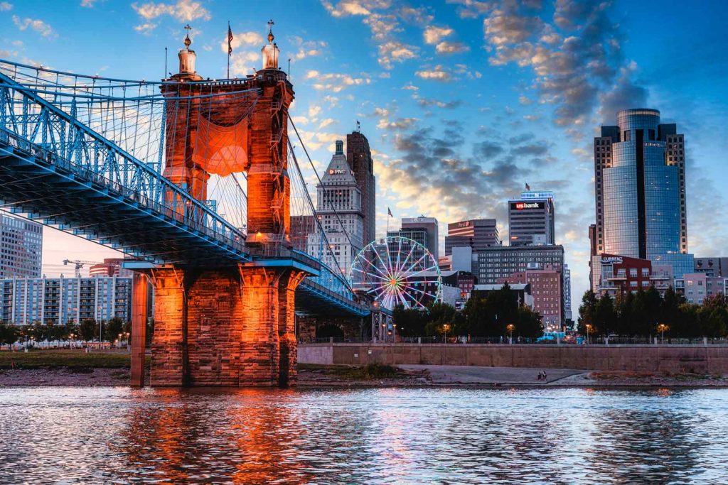 The Cincinnati skyline at sunset featuring the John A. Roebling Suspension Bridge, the Ohio River, and a ferris wheel.
