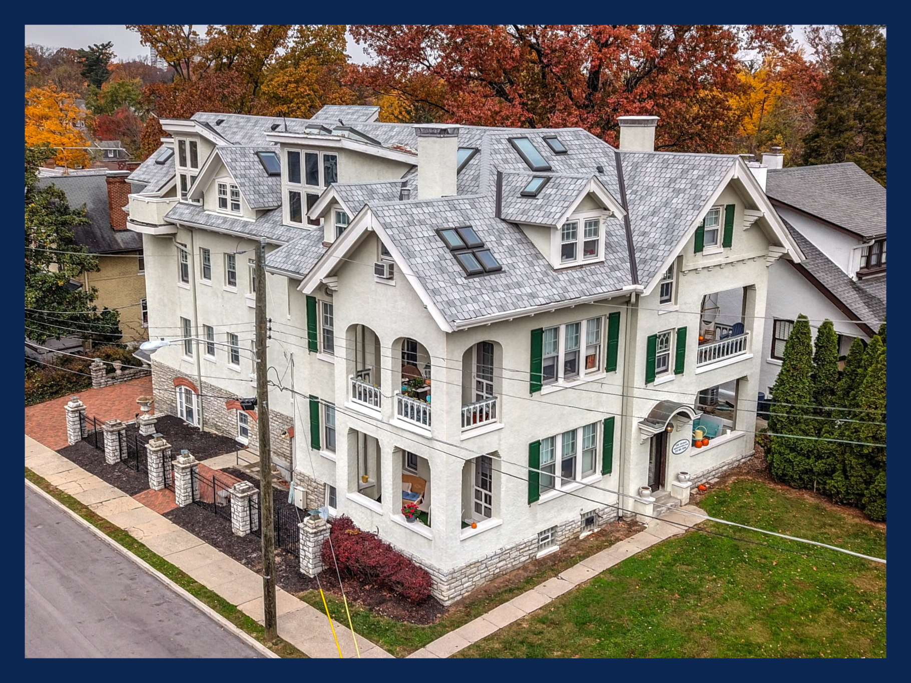 Large white multi-story house with green shutters, arched balconies, and a gray roof, surrounded by autumn trees and a black iron fence.