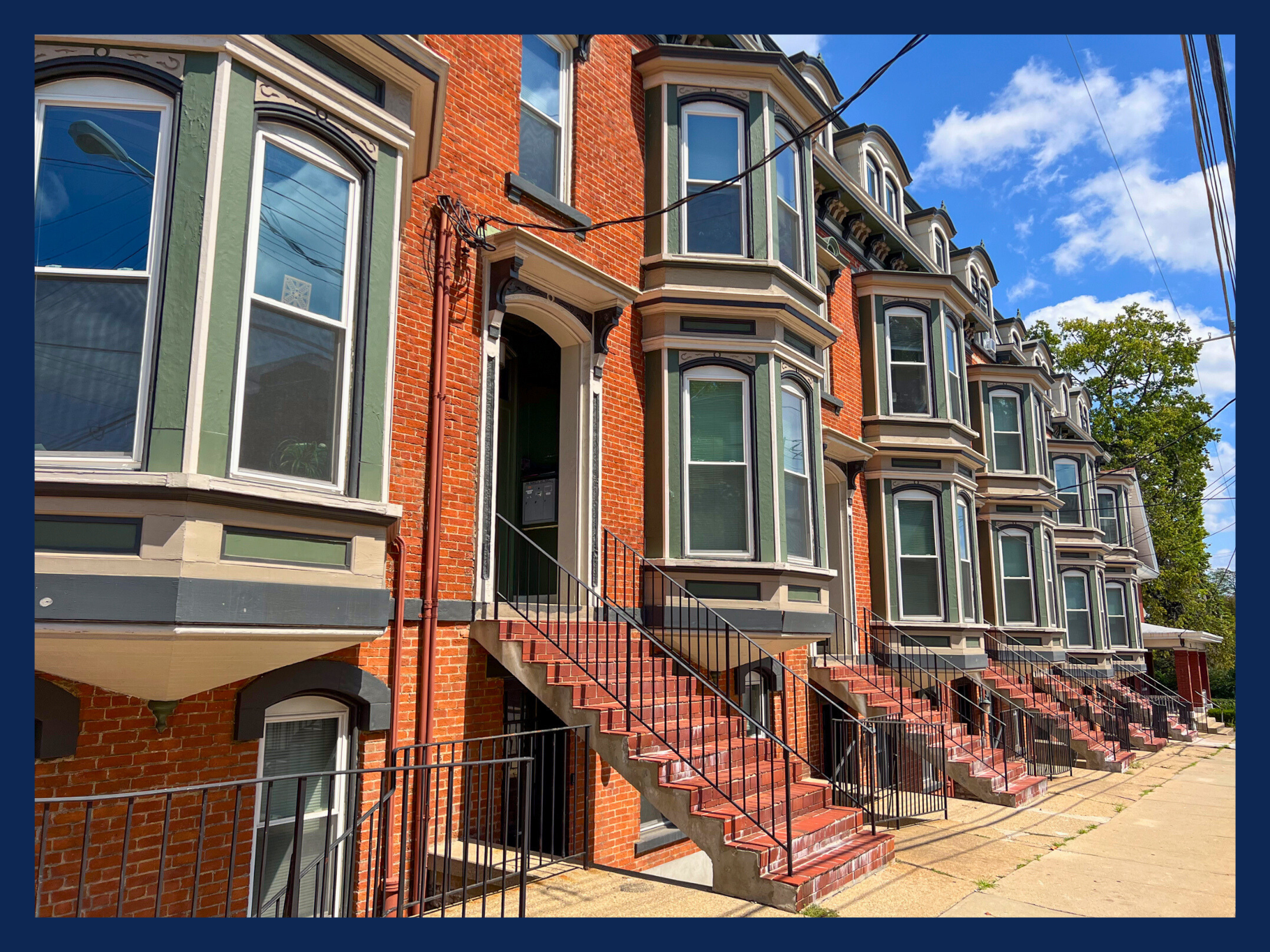 Row of historic red brick townhouses with bay windows and black iron railings, set against a bright blue sky with scattered clouds at 122 E McMilan Apartments.