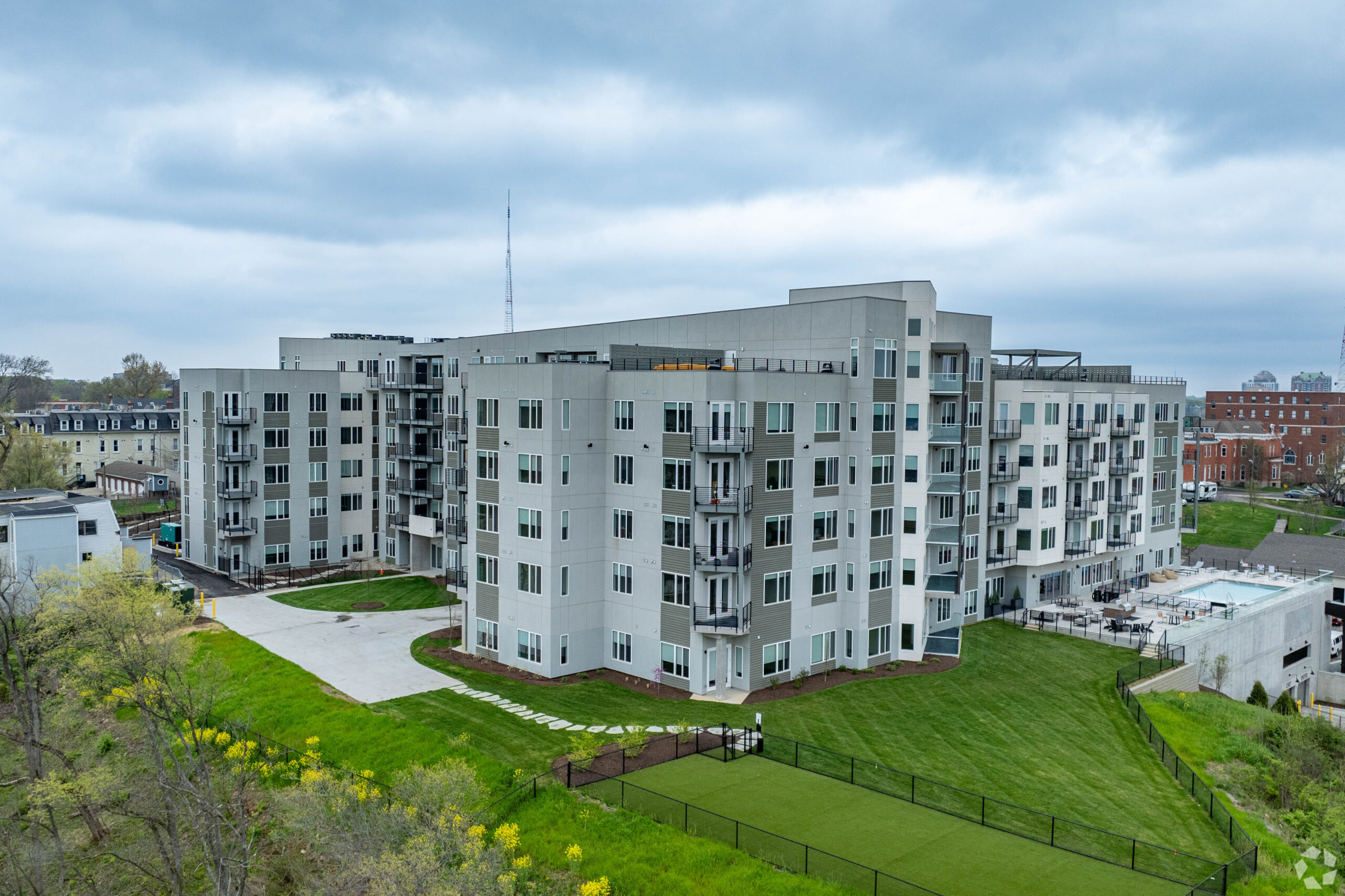 Modern multi-story apartment building at The Alto with balconies, large windows, green landscaping, and a fenced outdoor area under a cloudy sky.