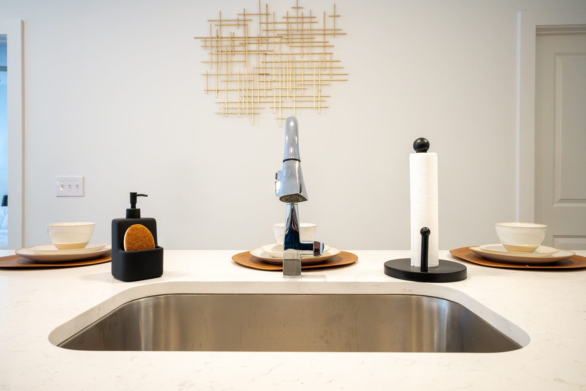 A stainless steel kitchen sink surrounded by quartz countertops in front of three place settings, a paper towel holder, and a soap dispenser at a breakfast bar.